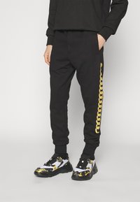 Černé bavlněné tepláky mají zlatý řetězový vzor na levé straně. Jsou kombinovány s černobílými teniskami se zlatými detaily.