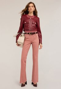 Giacca corta in pelle borgogna con due tasche frontali, abbinata a pantaloni a zampa d'elefante rosa chiaro e una borsa bianca. Sotto, un top in maglia testurizzata.
