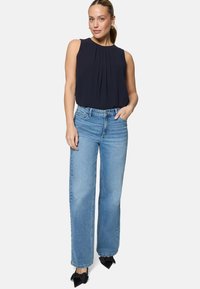 Femme avec queue de cheval portant un chemisier sans manches bleu marine, un jean large bleu clair et des chaussures noires pointues ornées de nœuds, debout avec une main dans la poche.