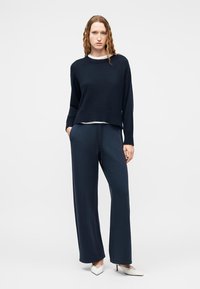 Γυναίκα με σγουρά μαλλιά που φορεί μαύρο πουλόβερ, μπλε παντελόνι με φαρδιά πόδια και λευκά μυτερά τακούνια, στέκεται με τα χέρια στις τσέπες.