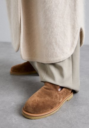 Pantofi slip-on din suede maro, cu vârf rotund, talpă din cauciuc și detalii cu decupaj lateral, asortați cu pantaloni bej deschis, ajustați.
