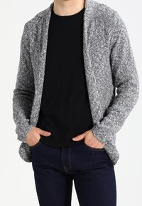 Cardigan grigio texture con una vestibilità rilassata, senza chiusure e con apertura frontale, indossato sopra una camicia nera e jeans scuri.