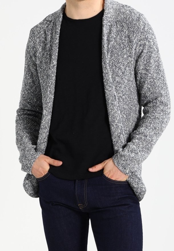 Cardigan grigio texture con una vestibilità rilassata, senza chiusure e con apertura frontale, indossato sopra una camicia nera e jeans scuri.
