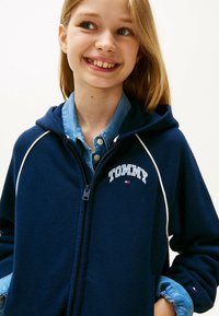 Granatowy hoodie na zamek z białymi akcentami i logo "TOMMY". Nałożony na jasnoniebieską koszulę z guzikami. Posiada kaptur i miękką fakturę.