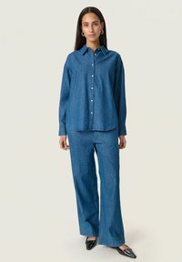 Set de cămașă din denim și pantaloni cu picioare largi în albastru mediu. Cămașa are un guler cu nasturi, mâneci lungi și o margine curbată; pantalonii sunt largi.