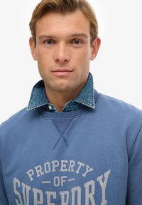 Sudadera azul con la inscripción "PROPERTY OF SUPERDRY" en gris claro, que presenta un cuello de mezclilla y puños acanalados, con textura suave y diseño casual.