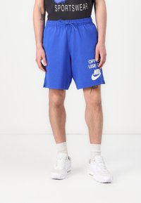 Calções Nike azuis com cintura elástica e cordão, apresentando o texto "USO OFICIAL APENAS" e o logótipo em branco. Usados com ténis brancos.