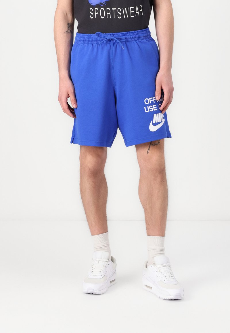 Calções Nike azuis com cintura elástica e cordão, apresentando o texto "USO OFICIAL APENAS" e o logótipo em branco. Usados com ténis brancos.