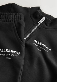 Melnas sporta krekls un bikses ar rāvējslēdzēju, ar balto "ALLSAINTS" logo un tekstu. Izgatavots no mīkstas, teksturētas auduma.