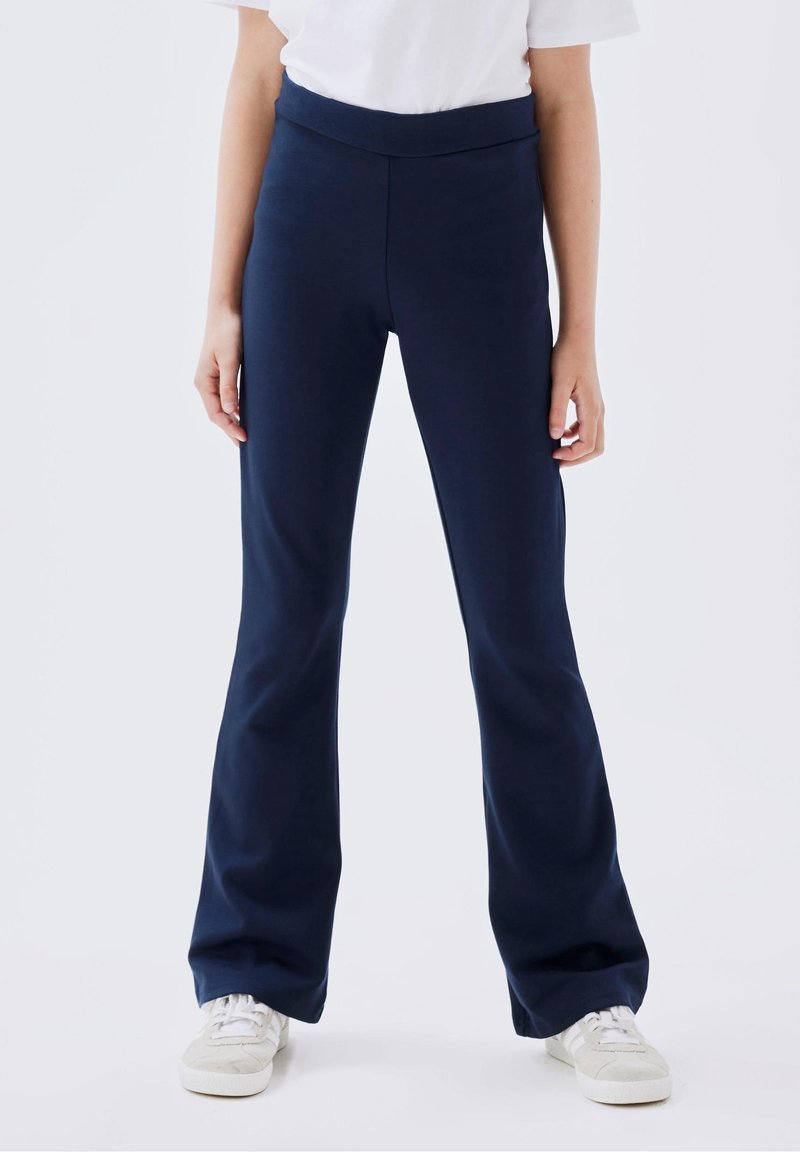 Personne portant un pantalon évasé taille haute bleu foncé, un haut blanc à manches courtes et des baskets blanches, debout devant un fond uni.
