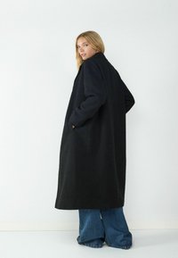 Lange zwarte jas van een gestructureerde stof, met twee zijzakken en een eenvoudige kraag. Gekozen boven een flared denim broek tegen een witte achtergrond.