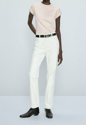 Person iført en lyserød top med korte ærmer, hvide bukser med høj talje og sort bælte samt sorte ankelstøvler mod en ensfarvet baggrund.