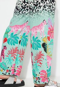 Ingezoomde afbeelding van een wijde broek in muntgroen met een jungleprint. Bevat roze tijgers, tropische bladeren en een verloop van zwart naar groen aan de bovenkant. Accenten van zilverkleurige hardware.