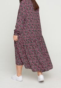 Robe maxi florale noire, ornée de fleurs roses et blanches. Manches longues, coupe ample et détail froncé à la taille. Portée avec des baskets blanches.