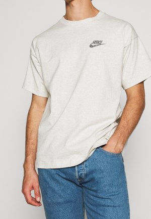 Mężczyzna w jasnobeżowej koszulce Nike z małym czarnym logo na piersi, w połączeniu z niebieskimi dżinsami, z ręką w kieszeni, stoi na jednolitym tle.