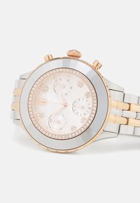 Orologio rotondo in acciaio inossidabile con bracciale bicolore argento e rosé. Presenta una lunetta decorata con cristalli e un quadrante rosa con texture.