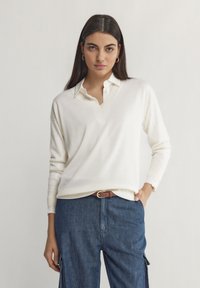 Cremefarbener V-Ausschnitt-Pullover mit langen Ärmeln, gestrickter Struktur, über einem weißen Hemd mit Kragen getragen. Kombiniert mit Jeans-Cargo-Hosen und einem braunen Gürtel.