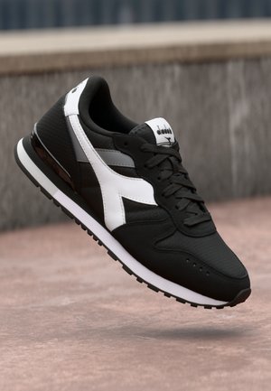 Chaussure de sport noire avec une tige texturée, des accents blancs et une semelle noire. Dotée d'un design à lacets et d'un logo sur la languette.