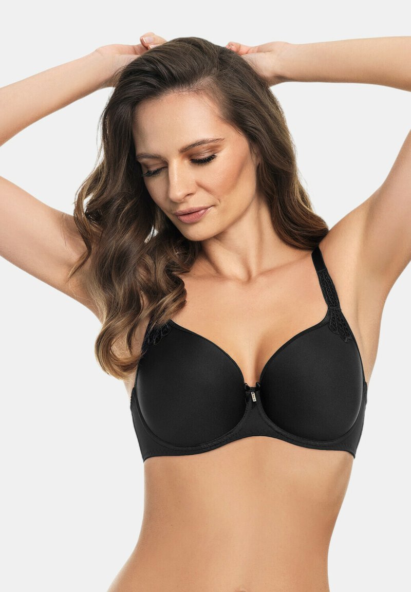 Donna che indossa un reggiseno imbottito nero con dettagli in pizzo sulle spalline, in posa con le braccia alzate e lo sguardo rivolto verso il basso su uno sfondo semplice.