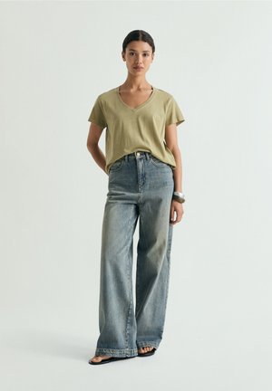 Giovane donna in piedi che indossa una maglietta verde oliva con scollo a V, jeans a gamba larga sbiaditi, sandali neri e un braccialetto d'argento al polso sinistro.
