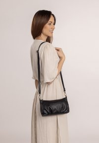 Zwarte leren handtas met een zachte, gerimpelde textuur, verstelbare riem en een eenvoudige ritssluiting. Draagstijl als crossbody.