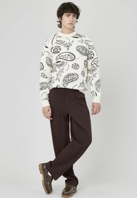 Pull blanc avec motif cachemire noir, pantalon marron et chaussures marron avec détail en chaîne. Le modèle se tient avec une main sur la hanche.