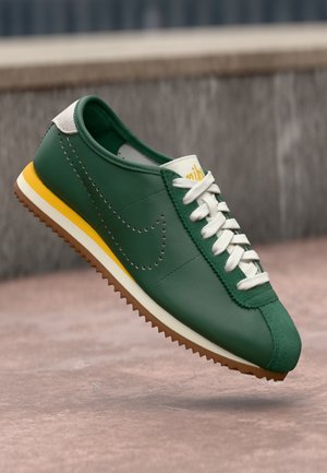 Green leather sneaker with white laces, perforated swoosh logo, yellow stripe on midsole, and brown rubber outsole suspended above ground.