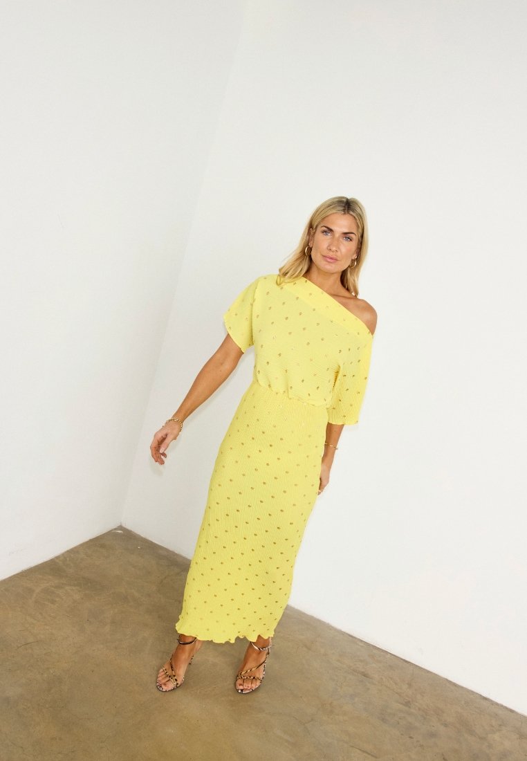 Femme portant une robe midi jaune à une épaule avec de petits pois, debout pieds nus sur un sol en béton contre un mur blanc.