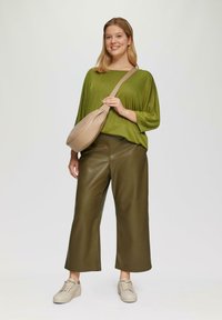 Vrouw die glimlacht, gekleed in een olijfgroene top, brede leren broek, beige sneakers en een beige schoudertas, op een witte achtergrond.