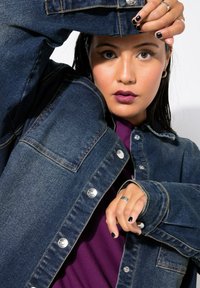 Chaqueta de mezclilla en azul oscuro con botones a presión plateados, bolsillos prominentes y un ajuste ligeramente holgado, combinada con una blusa morada.