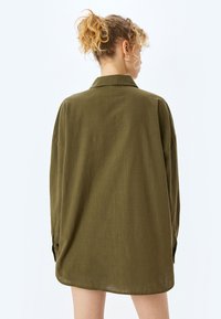 Camisa de manga larga color verde oliva con cuello y corte relajado; incluye puños abotonados y un dobladillo ligeramente curvado para un aspecto casual.