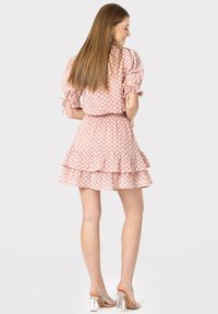 Rochie roz cu mâneci bufante, cu buline albe, tiv cu volane în straturi, talie elastică și accente cu funde pe mâneci. Purtată cu sandale cu toc transparent.