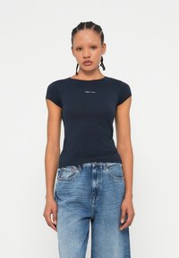 Femeie cu părul împletit, îmbrăcată cu un tricou ajustat de culoare albastru închis cu mâneci scurte și blugi lejeri de culoare albastru deschis, stând în fața unui fundal simplu.