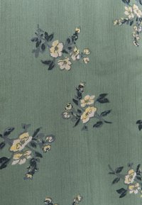 Kankaita kukkakuviolla vihreällä pehmeällä tekstuurilla; siinä on keltaisia ja vaalean valkoisia kukkia tummilla lehdillä, luoden hienovaraisen muotoilun.