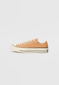 Canvas sneakers i lys orange med hvite snorer og gummitåhette. Har en svart stripe langs sålen. Lav profildesign.