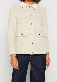 Chaqueta de pana beige con cuello de piel sintética, botones a presión plateados y cuatro bolsillos frontales. Presenta un acabado de tela texturizada.