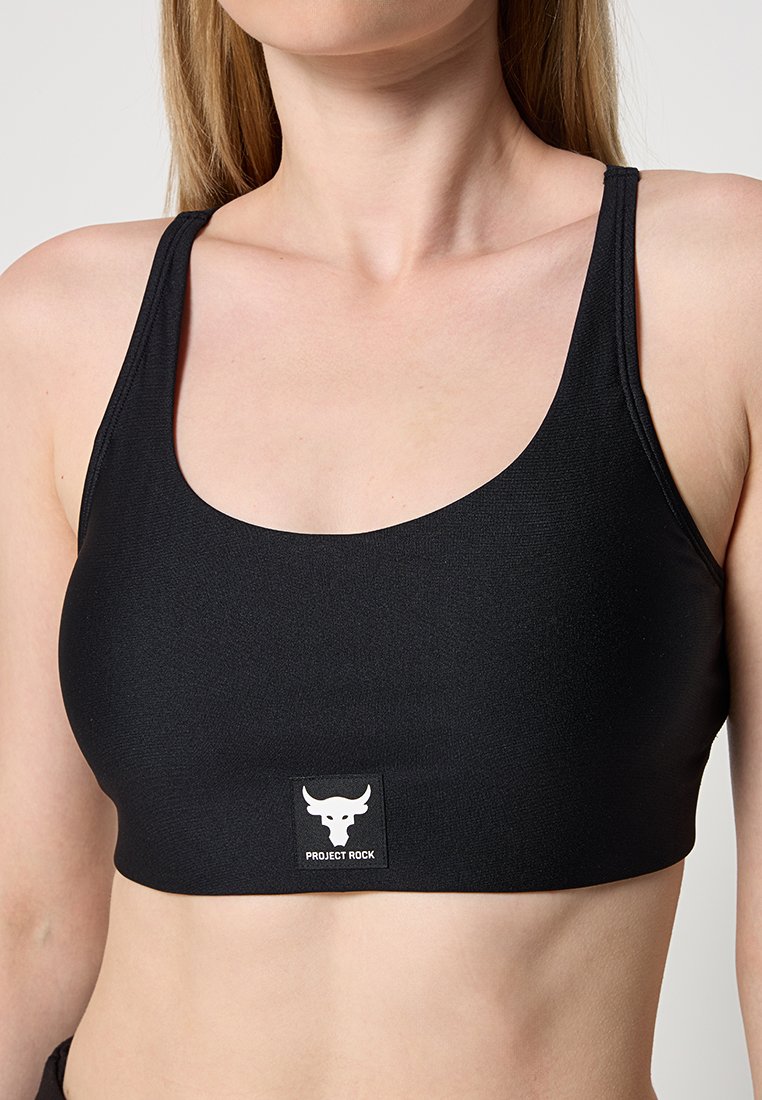 Sujetador deportivo negro con escote en forma de barco, hecho de un material suave y elástico. Presenta un parche de logo blanco que dice "PROJECT ROCK".