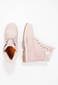 Une paire de bottes en suede rose clair à lacets, avec un col de cheville texturé et une épaisse semelle en caoutchouc, présentée en vue de dessus et de côté sur fond blanc.