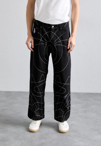 Pantalons larges noirs en denim avec des motifs de lignes abstraites blanches, dotés de poches avant et d'un épinglette décorative sur la taille.