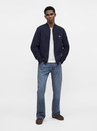 Homme debout, les mains dans les poches, portant un blouson bomber bleu marine, un t-shirt blanc, un jean bleu et des chaussures brunes sur un fond blanc uni.