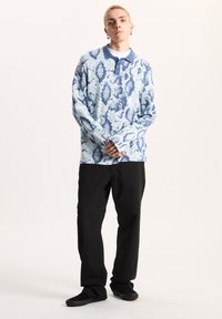 Ljusblå långärmad pikétröja med ett strukturerat ormmönster, som har en kontrasterande krage och knappar, tillsammans med svarta byxor och skor.