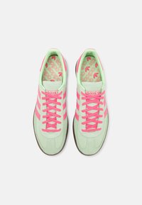 Zelené semišové tenisky s růžovými akcenty a tkaničkami. Disponují třemi růžovými pruhy a vzorovanou vnitřní stélkou. Zaoblená špička a hnědá gumová podrážka.