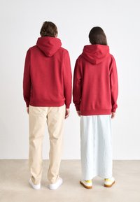 Sudaderas rojas con capuchas ajustables, llevadas por dos figuras. Una viste pantalones color beige y zapatillas blancas, y la otra un falda azul clara con acentos amarillos.