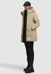 Beige pufferjas met een capuchon, zwarte rits en logo patch. Zwarte leggings en veterschoenen in zwart voltooien de outfit.