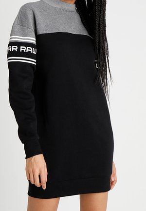 Robe-pull noire et grise à manches longues avec des rayures blanches et le texte "G-STAR RAW" sur le haut du bras, portée par une personne aux cheveux tressés.