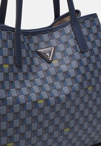 Niebieska torebka ze wzorem, wykonana z trwałego materiału, z trójkątną plakietką z logo oraz usztywnioną konstrukcją z teksturowanymi akcentami.