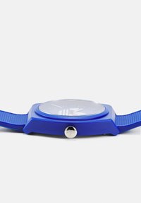 Montre en silicone bleue avec un bracelet texturé, un boîtier carré, une couronne argentée et un cadran rond avec des marquages horaires et des aiguilles blanches.