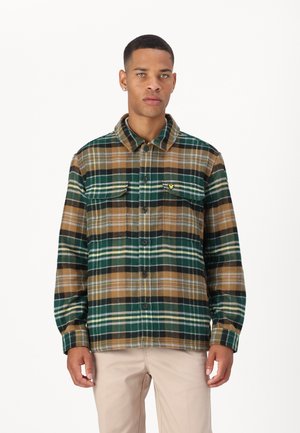 Plaid flannel shirt in green, brown, and cream with two chest pockets, button closure, and a small yellow emblem on the left side.