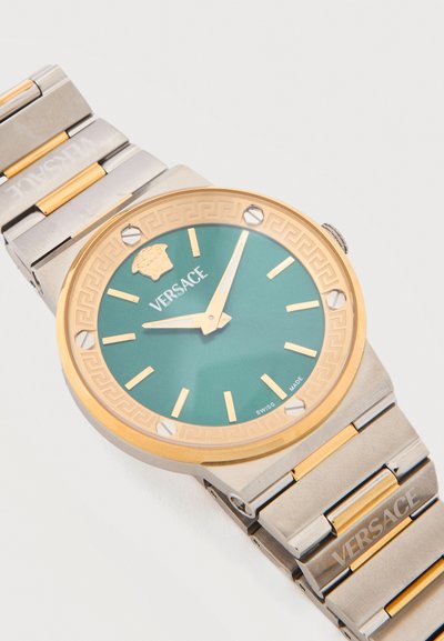 Náramkové hodinky Versace so zlatým a strieborným kovovým remienkom, zeleným ciferníkom, zlatými značkami a logom Medusa na pozícii 12 hodín, zobrazujúce čas 10:07.