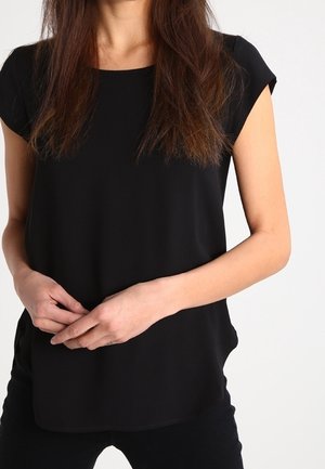 Vrouw draagt een eenvoudige zwarte korte mouwen blouse, handen losjes in de taille gevouwen, met lang bruin haar over de schouders.